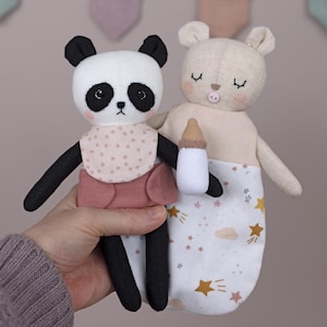 以下が含まれることがあります： 黒と白のパンダとベージュのクマのぬいぐるみ2体が、白いボトルを持っています。パンダはピンクと白のドット柄のスタイとピンクのスカートを着ています。クマは白と金の星柄の服を着ています。