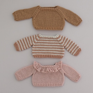 May include: Three knitted sweaters for dolls. The top sweater is brown, the middle sweater is striped in white and brown, and the bottom sweater is pink with a ruffled neckline.