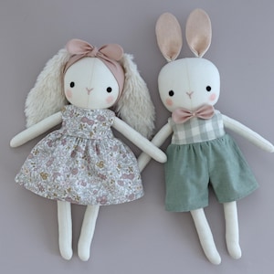 May include: Two handmade plush bunny dolls, one wearing a floral dress and the other wearing a green and white gingham overall with a pink bow tie.