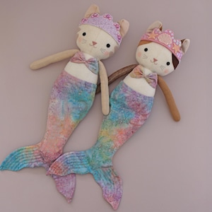 May include: Two handmade mermaid cat dolls. Each doll has a cream-colored face with embroidered features, a bow tie, and a colorful mermaid tail. One wears a purple crown, the other a pink crown.