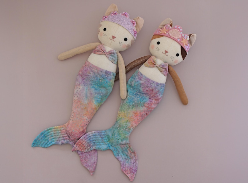 May include: Two handmade mermaid cat dolls. Each doll has a cream-colored face with embroidered features, a bow tie, and a colorful mermaid tail. One wears a purple crown, the other a pink crown.