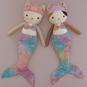 May include: Two plush mermaid cat dolls. Each doll has a cream-colored face with cat features, a pink bow, and a mermaid tail with blue, pink, and yellow tie-dye patterns. They wear jeweled crowns and hold hands.