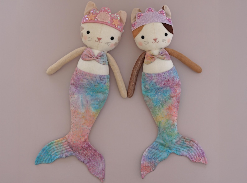 May include: Two plush mermaid cat dolls. Each doll has a cream-colored face with cat features, a pink bow, and a mermaid tail with blue, pink, and yellow tie-dye patterns. They wear jeweled crowns and hold hands.