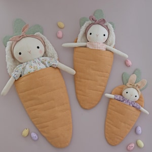 May include: Three plush bunny dolls in carrot-shaped sleeping bags. The bunnies have soft white fur, pink bows, and wear floral or gingham patterned clothing. The carrot beds are orange with green tops, and pastel Easter eggs are scattered around.
