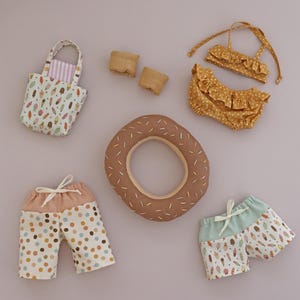 Puede incluir: Colección de accesorios de playa para muñecas, que incluye un bikini amarillo con lunares, dos pares de pantalones cortos con diferentes estampados, una boya con forma de rosquilla, un bolso de mano con estampado de helados y dos pequeños bloques de madera. Los artículos están dispuestos sobre una superficie de color lila claro.