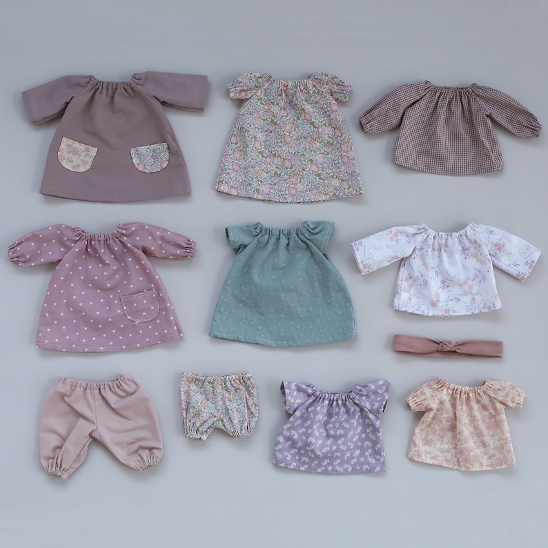 K&ouml;nnte beinhalten: Eine Sammlung von zehn Puppenkleidern, darunter Kleider, Hemden, Hosen und ein Stirnband. Die Kleidung ist aus verschiedenen Stoffen gefertigt und hat unterschiedliche Muster, darunter Punkte, Blumenmuster und Gingham.