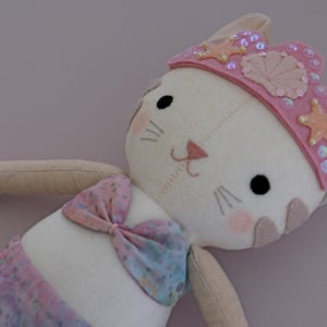 May include: A handmade plush cat doll dressed as a mermaid. The doll has a white body, pink cheeks, and a pink crown with sequins and starfish accents. It wears a seashell-patterned bikini top and a purple and pink mermaid tail.