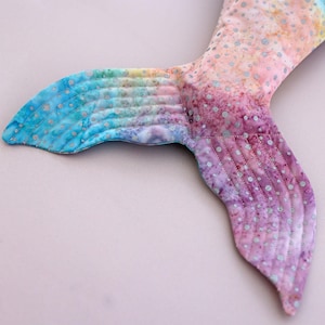 May include: A colorful mermaid tail in shades of blue, yellow, pink, and purple. The fabric is quilted with a dotted pattern. The tail is designed with a curved shape and is likely a decorative item or part of a costume.