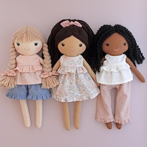 May include: Three fabric dolls with yarn hair and embroidered facial features. The left doll has blonde braided hair, a peach gingham top, and blue shorts. The center doll has dark brown hair with a bow and a floral dress. The right doll has black curly hair, a white eyelet top, and pink gingham pants.