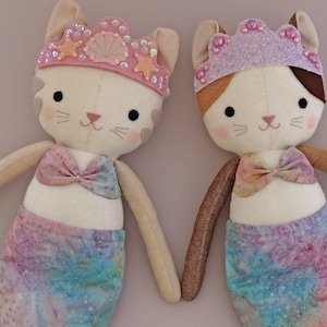 May include: Two handmade cat mermaid dolls. Each doll has a cream-colored face with black eyes, pink cheeks, and a pink nose. They wear a crown and a bow. The mermaid tails are a watercolor blend of blue, pink, and purple.