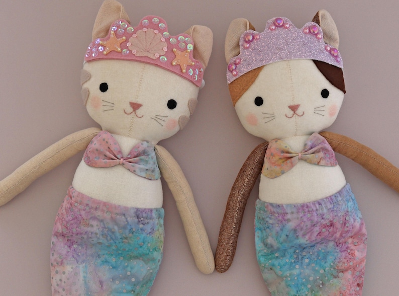 May include: Two handmade cat mermaid dolls. Each doll has a cream-colored face with black eyes, pink cheeks, and a pink nose. They wear a crown and a bow. The mermaid tails are a watercolor blend of blue, pink, and purple.