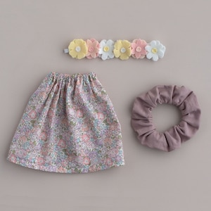 Puede incluir: Una falda de estampado floral con cintura elástica, una scrunchie morada y una diadema de flores de fieltro amarillo, rosa y blanco.