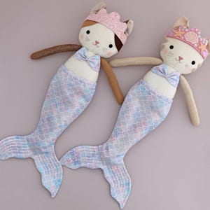 May include: Two plush mermaid cat dolls. Each doll has a cat face with a crown, a bow, and a mermaid tail. The tails are light blue, pink, and purple. The dolls have brown arms and are lying on a light purple surface.