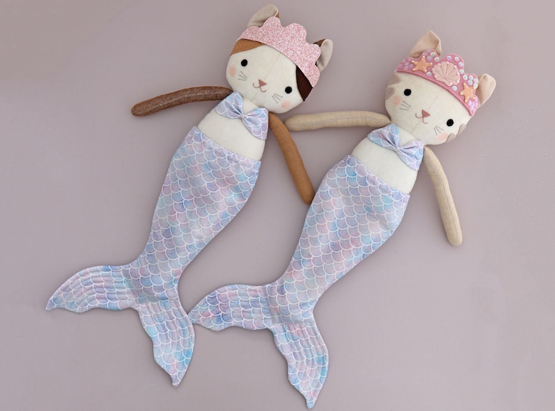 May include: Two plush mermaid cat dolls. Each doll has a cat face with a crown, a bow, and a mermaid tail. The tails are light blue, pink, and purple. The dolls have brown arms and are lying on a light purple surface.