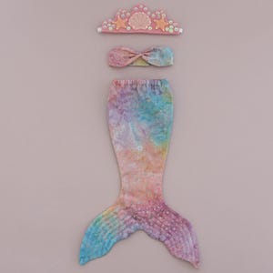May include: A mermaid costume set, including a tail, top, and crown. The tail is a gradient of blue, purple, and pink. The top is a matching fabric, and the crown is pink with star and seashell accents. A whimsical outfit for dress-up.