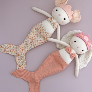 May include: Two plush mermaid dolls with bunny faces. One doll has a floral print tail and the other has a pink scale-like tail. Both dolls wear pink crowns and bows. The dolls are on a lavender background.
