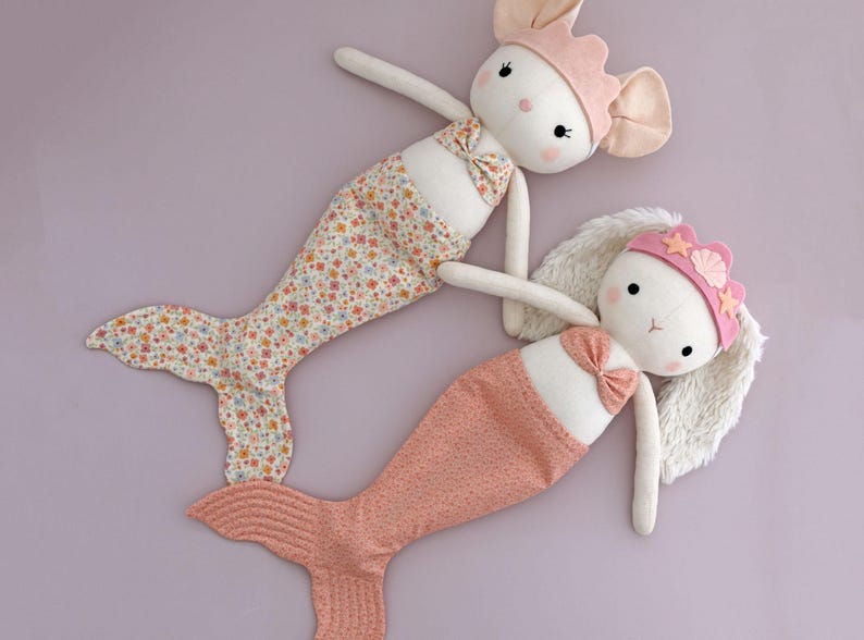 May include: Two plush mermaid dolls with bunny faces. One doll has a floral print tail and the other has a pink scale-like tail. Both dolls wear pink crowns and bows. The dolls are on a lavender background.