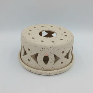 Può includere: Un scaldacandele in ceramica beige macchiato con un design circolare. Lo scaldacandele ha forme ritagliate e fori per la ventilazione. La base è di colore beige macchiato abbinato.