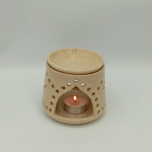 May include: A white ceramic oil diffuser with a small candle burning inside. The diffuser has a decorative design with holes around the top and bottom.