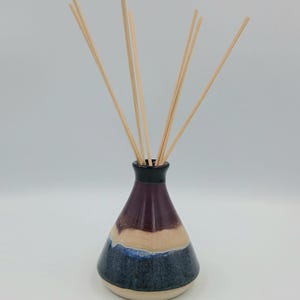 Może przedstawiać: Ceramiczny dyfuzor olejków eterycznych z warstwowym wzorem w odcieniach niebieskiego, beżowego i bordowego. Dyfuzor ma czarną obwódkę i zawiera kilka drewnianych patyczków do rozprowadzania zapachu. Jest to dekoracyjny element zapachowy do domu.