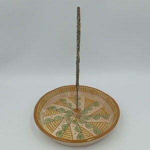 May include: A ceramic incense burner with a circular design featuring green and yellow geometric patterns. A single incense stick is placed in the center of the burner.