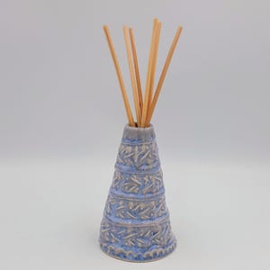 Peut inclure: Un diffuseur en céramique de forme conique avec un émail texturé bleu clair et gris, orné de bandes horizontales répétitives de motifs sculptés en forme de feuilles. Cinq fins bâtonnets en bois clair sont insérés dans l'ouverture étroite.