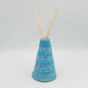 May include: A conical ceramic reed diffuser vessel in a vibrant turquoise blue glaze, featuring a repeating embossed floral pattern across its surface and containing five thin, light-colored wooden reeds extending from the narrow top opening.