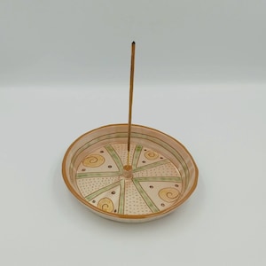Può includere: Un bruciatore di incenso in ceramica con un design decorativo in tonalità di marrone, verde e giallo. Il bruciatore ha un foro centrale per un bastoncino di incenso, che è incluso.