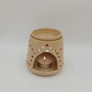 Può includere: Un diffusore di oli essenziali in ceramica beige con una candela all'interno. Il diffusore ha una parte superiore rotonda e una base con fori decorativi. La fiamma della candela è visibile attraverso l'apertura. Per aromaterapia.
