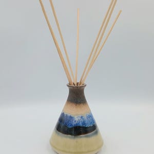 Op de afbeelding: Een keramische rietverspreider met een conische vorm en een smalle hals. De vaas heeft kleurstroken, waaronder beige, blauw en bruin. Verschillende houten rietjes zijn in de opening gestoken, klaar om geur te verspreiden.