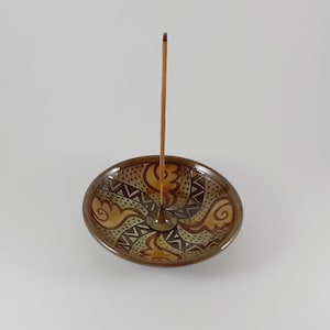 Può includere: Un porta incenso in ceramica marrone con un design geometrico. Il porta incenso ha un piccolo foro al centro per un bastoncino di incenso. Un bastoncino di incenso è in piedi nel porta incenso.