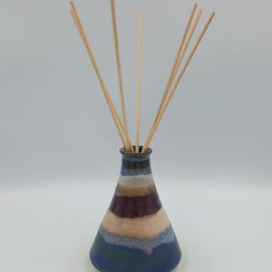 Op de afbeelding: Een keramische etherische olie diffuser met een conische vorm en een gestreept patroon in tinten blauw, bruin en paars. De diffuser bevat verschillende houten rietjes die omhoog steken. De basis is donkerblauw.