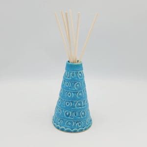Op de afbeelding: Een conische keramische geurverspreider met een helder turquoise blauwe glazuurlaag. De buitenkant is voorzien van een herhalend reliëfpatroon van cirkels en horizontale lijnen. Vijf lichtgekleurde houten stokjes steken uit de smalle opening.