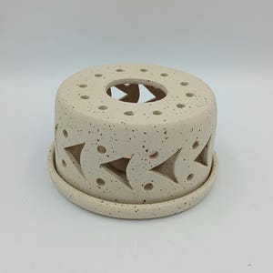 Peut inclure: Un chauffe-bougie en céramique beige moucheté de forme circulaire. Le chauffe-bougie présente des découpes en forme de flèche sur les côtés et de petits trous sur le dessus. Il repose sur une base assortie, créant une pièce décorative et fonctionnelle.