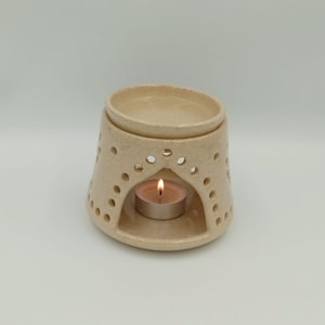 May include: A ceramic essential oil diffuser with a white glaze and a decorative cutout design. A small tea light candle is placed in the centre of the diffuser.