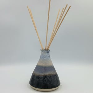 Op de afbeelding: Een keramische etherische olie diffuser met een gradiëntontwerp. De vaas loopt over van een lichtblauwe bovenkant naar een beige, dan een donkerblauwe en tenslotte een zwarte basis. Er zijn verschillende houten rietjes in de vaas gestoken.