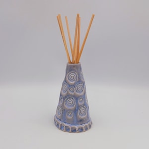 May include: A cone-shaped ceramic reed diffuser with a light blue and grey glaze. The surface features embossed spiral patterns and a scalloped base. Five thin, light brown wooden reeds are inserted into the narrow opening at the top.
