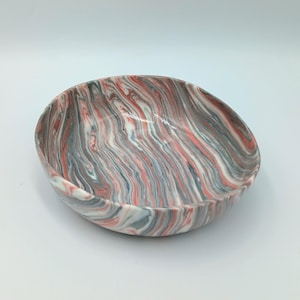 May include: A shallow, round bowl featuring a marbled pattern with swirling streaks of white, coral pink, and slate grey. The surface has a smooth, glossy finish, and the design creates a continuous, fluid appearance across the entire exterior.