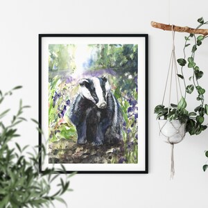 May include: Watercolor painting of two badgers in a forest setting, framed in a black border. The badgers are depicted with black and white fur, surrounded by green foliage and purple flowers. The artwork is displayed on a white wall.