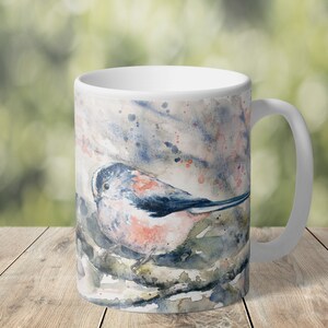 May include: White ceramic mug with a watercolor illustration of a blue and pink bird perched on a branch. The background is a soft blue and pink watercolor wash.