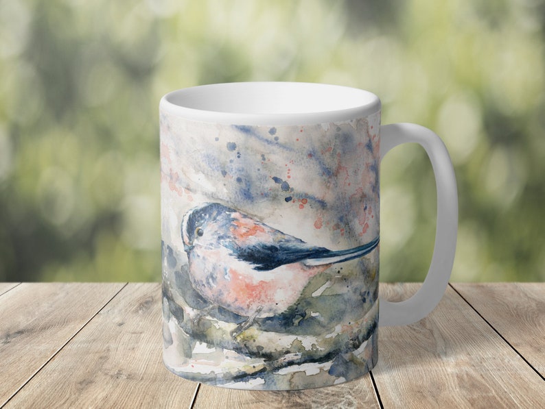 May include: White ceramic mug with a watercolor illustration of a blue and pink bird perched on a branch. The background is a soft blue and pink watercolor wash.