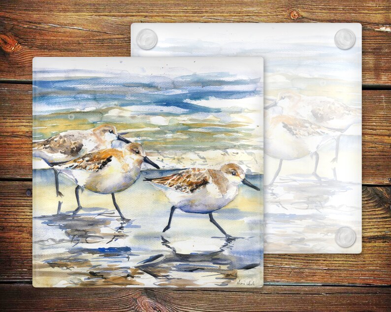 May include: Three sandpipers, painted in watercolor, stand on a sandy beach with blue water in the background. The birds are brown, white, and gray with long legs and beaks. The painting is on a glass coaster.