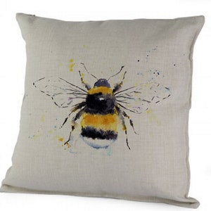 Puede incluir: Una almohada de lino beige con una pintura de acuarela de un abejorro. El abejorro es amarillo y negro con alas transl&uacute;cidas. El fondo es beige claro con salpicaduras de acuarela amarilla y azul.