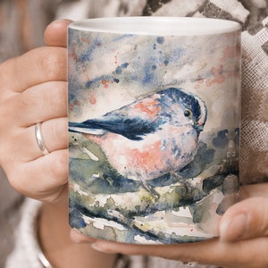 May include: A white ceramic mug with a watercolor design of a blue and pink bird perched on a branch. The background is a soft blue and gray with hints of pink and green.