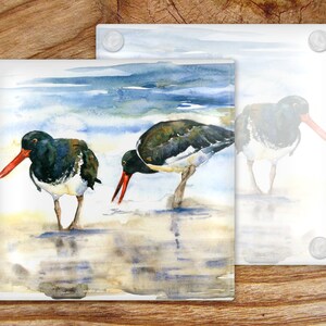 May include: Two black and white oystercatchers with red beaks stand in shallow water on a sandy beach. The background is a watercolor painting of blue water and a cloudy sky.