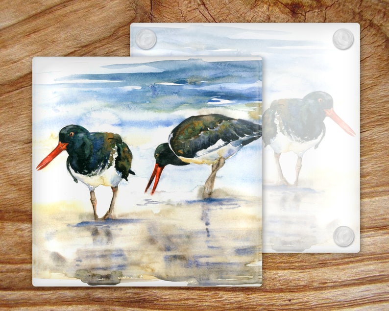 May include: Two black and white oystercatchers with red beaks stand in shallow water on a sandy beach. The background is a watercolor painting of blue water and a cloudy sky.