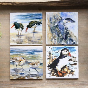 May include: Set of four square ceramic coasters featuring watercolor paintings of different birds. The coasters depict a pair of oystercatchers, a puffin, a seagull, and a group of sandpipers.