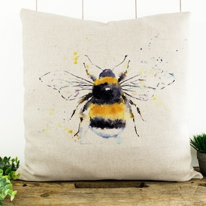 Puede incluir: Una pintura de acuarela de un abejorro sobre una almohada de lino beige. El abejorro es amarillo y negro con alas transl&uacute;cidas. El fondo es beige claro con salpicaduras de acuarela amarilla y azul.