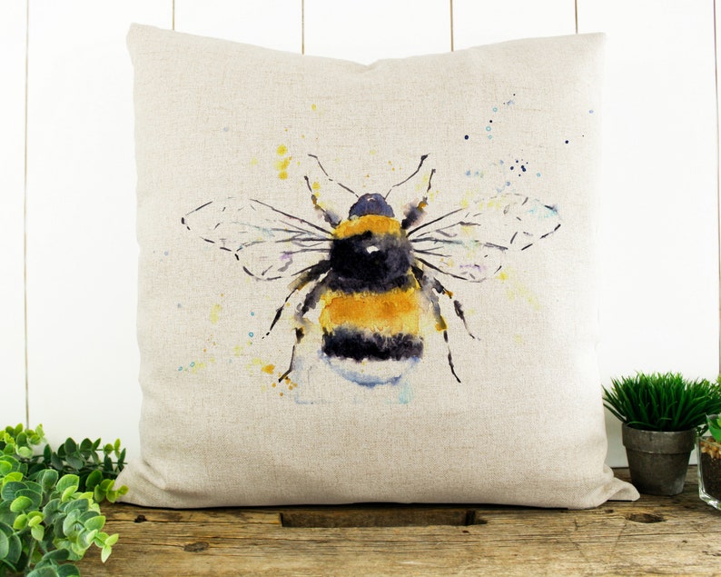 Puede incluir: Una pintura de acuarela de un abejorro sobre una almohada de lino beige. El abejorro es amarillo y negro con alas transl&uacute;cidas. El fondo es beige claro con salpicaduras de acuarela amarilla y azul.