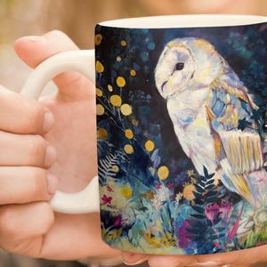 May include: White ceramic mug featuring a detailed painting of a barn owl. The owl is depicted with white, blue, and gold feathers, set against a dark background with colorful floral accents. The mug has a white handle.